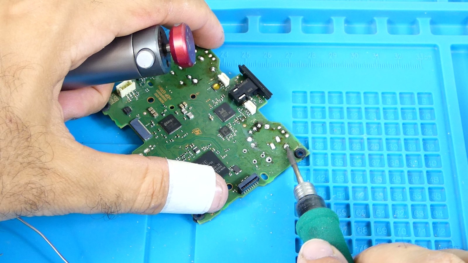 Técnico sosteniendo simultáneamente una bomba de succión desoldadora y un soldador de precisión sobre la placa base de un mando DualSense, trabajando un pin con estaño fundido